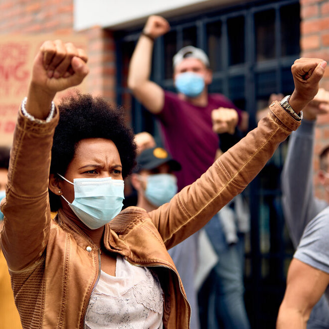 Black woman with raised fists wearing protective face mask while supporting anti-racism demonstrations.