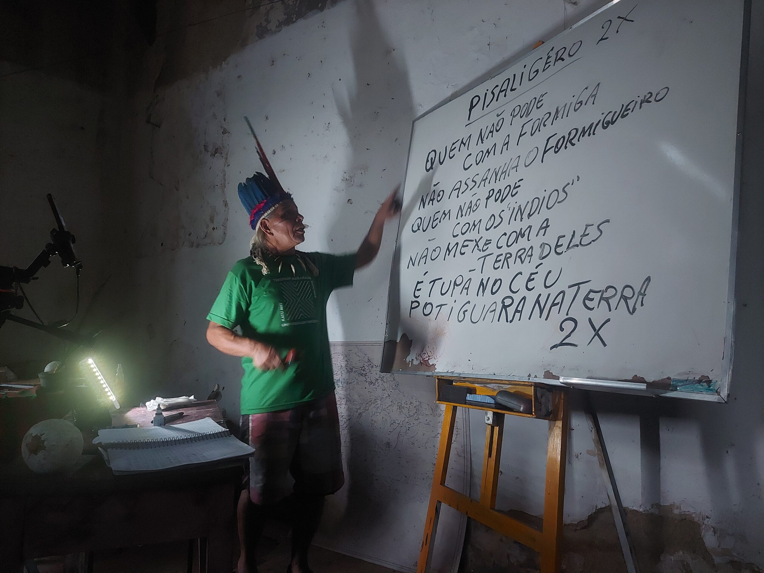 Chief José Urutau is standing in a classroom facing a whiteboard with writing on it and gesturing towards it