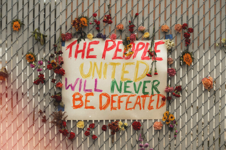 A handwritten sign on a chain-link fence reads "The People United Will Never Be Defeated" in rainbow-colored letters, surrounded by dozens of flowers tucked into the fence links.